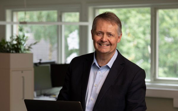 A man with a black jacket and a blue shirt is smiling at the camera. A laptop is open on the table in front of him. Behind him there are windows with lush green trees outside.