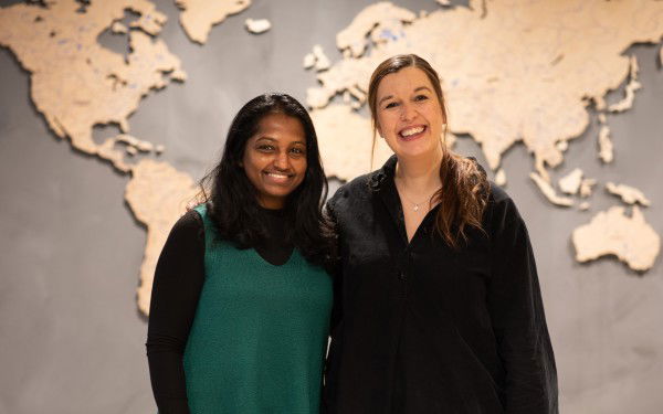 To women are smiling in front of a grey wall with a world map.