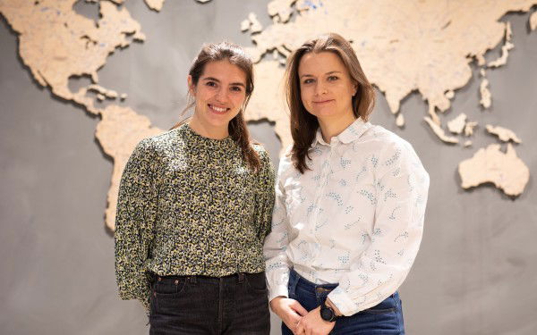 Two women are standing in front of grey wall with a beige world map.